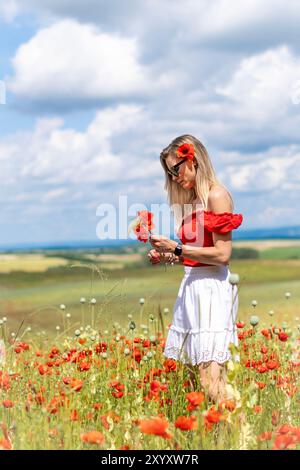 Blonde junge Frau in Sonnenbrille mit Mohn im Haar, die Blumen auf einem Feld aus rotem Mohn pflückt. Stockfoto