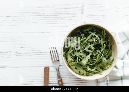 Grüne frische Rucola-Salatblätter in Schüssel, Gabel und Messer auf weißem hölzernem Hintergrund, weißes Handtuch. Konzept für gesunde Ernährung. Stockfoto