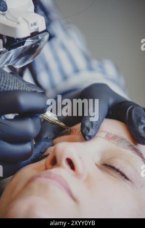 Die Kosmetikerin tätowiert sorgfältig das dauerhafte Augenbrauen-Make-up auf eine Frau, trägt schwarze Handschuhe zur Hygiene. Die Frau legt sich hin, die Augen geschlossen, vertraut der Kunst Stockfoto