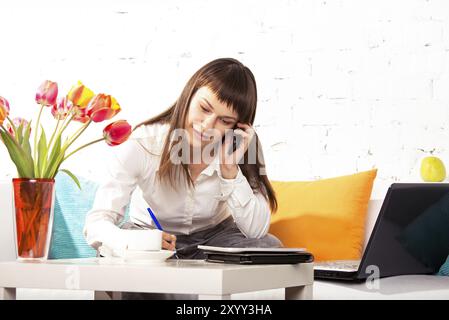 Junge brünette Frau, die zu Hause telefoniert und am Laptop arbeitet Stockfoto