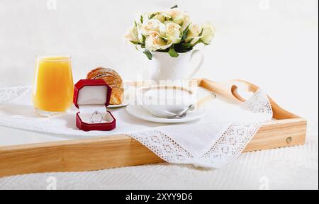 Romantisches Frühstück im Bad mit Croissant und einem Glas Orangensaft und einer Tasse Kaffee auf Holztablett mit Verlobungsring in roter Box und Bouquet Stockfoto