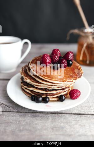 Saftige Pfannkuchen mit Beeren und Honig auf einem weißen Teller, Löffel, Glas, Holztisch, Kaffeetasse. Stockfoto