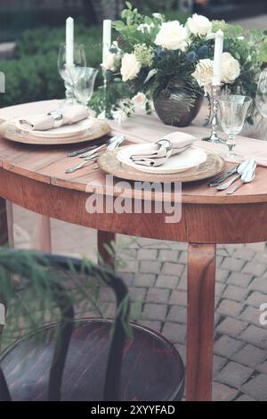 Kerzen und Vase mit weißen Rosen auf einem runden Tisch in der Nähe von Weingläsern während der Hochzeit im Garten Stockfoto