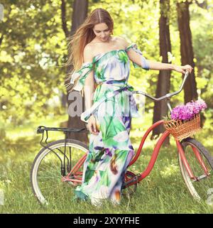 Schöne blonde Frau, die ein schönes Kleid trägt und Spaß im Park hat, mit dem Fahrrad, das einen schönen Korb voller Pfingstrosen-Blumen trägt. Vintage-Landschaft. Schön Stockfoto