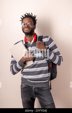 Lächelnder junger Afroamerikaner mit Brille und Dreadlocks, der Notebook hält und Rucksack trägt. Porträt eines selbstbewussten Studenten in Casua Stockfoto