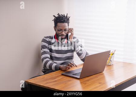 Junger afroamerikanischer Profi in lässigem gestreiftem Pullover, der von zu Hause aus arbeitet. Mann mit Dreadlocks und Brille, der Laptop und Smartphone für Busin benutzt Stockfoto