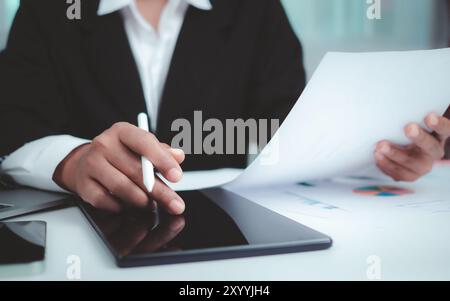 Geschäftsfrau in schwarzem Anzug, die an Laptop-Computer, Papierdokumenten, Tablet und Rechner arbeitet, im Büro mit einem dunklen Hintergrund das Tablet mit den Händen berührt, Stockfoto