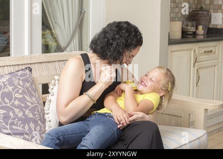 Eine junge Mutter und ihre Tochter spielen und haben Spaß, während sie draußen auf der Veranda ihres Hauses auf einer Couch sitzen Stockfoto