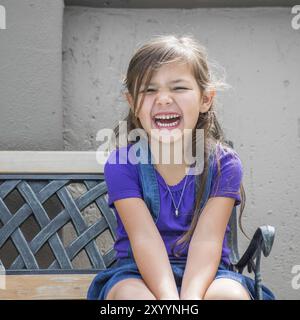 Das kleine Mädchen sitzt lachend auf der Außenseite der Bank Stockfoto