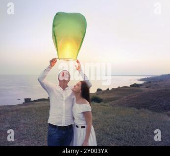 Junges Paar starten eine grüne chinesische Himmelslaterne in der Abenddämmerung am Meer Stockfoto