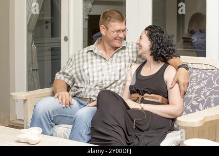 Ein junges Paar sitzt auf einer Bank auf der Terrasse, der Mann hat den Arm um die Frauen, den Hals, sie sehen sich an, während sie glücklich lächeln und lachen Stockfoto