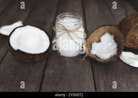 Gebrochene Kokosnüsse auf grauem hölzernem Hintergrund mit einem Glas rohen Bio extra nativen Kokosöl. Weißes Kokosnussfleisch. Stockfoto