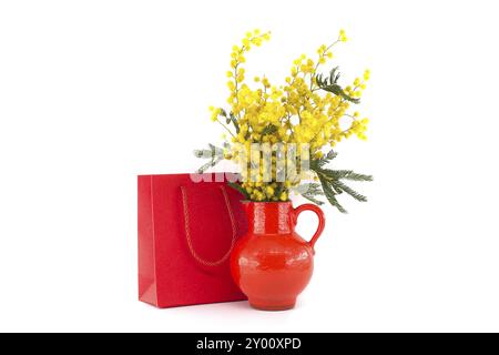 Mimosa Blumenstrauß in roter Vase in der Nähe von roter Geschenktüte isoliert auf weißem Hintergrund, Ostern, 8. März Frauentag Konzept Stockfoto