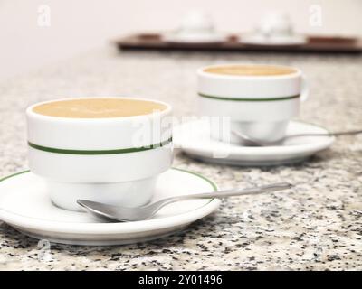 Cappuccino in zwei weißen Tassen, mit grünem Ring, nicht ausgerichtet, auf Steintisch, weiße Teller, mit Teelöffel an der Seite Stockfoto