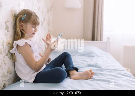 Ein kleines Mädchen sitzt im stilvollen Schlafzimmer auf dem Bett, hält das Telefon und führt per Smartphone einen Videoanruf, wobei es mit der Hand winkt. Kommunikation oder Homeschool Stockfoto