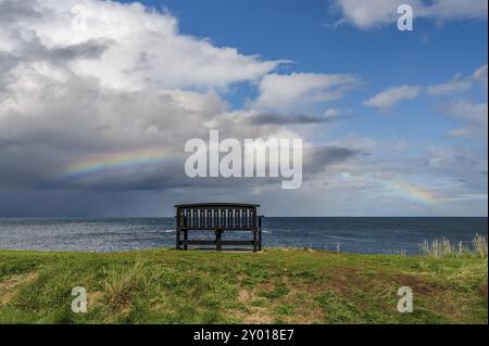 Eine Bank mit einem Regenbogen über der Nordsee Küste, im Benthall, Northumberland, England, UK gesehen Stockfoto