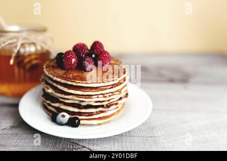 Saftige Pfannkuchen mit Beeren und Honig auf einem weißen Teller, Löffel, Glas, Holztisch. Stockfoto