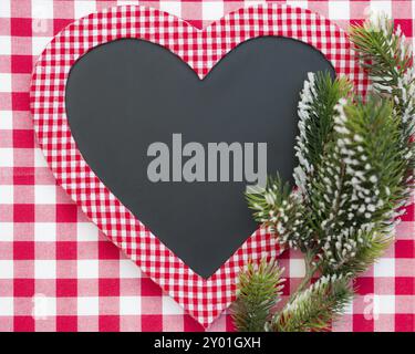 Karteikarte in Herzform mit Tannenbaumzweig auf roter Gingham-Tischdecke. Konzept für Winterferien Stockfoto