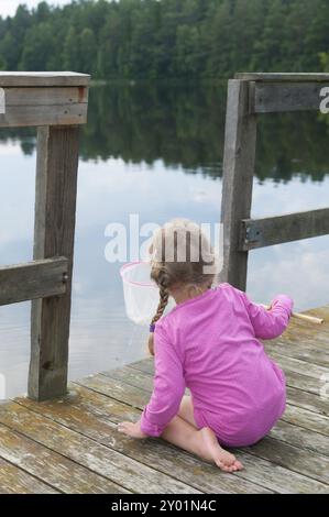 Niedliches kleines Mädchen kniet auf den Holzplanken eines Piers und fischt mit einem Landenetz im Licht eines skandinavischen Sommerabends Stockfoto