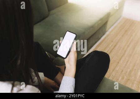 Kommunikationskonzept mit schwarzem Hear-Girl, das auf dem grünen Sofa sitzt und das Smartphone in der Hand hält, mit leerem weißen Bildschirm und Sonnenstrahl auf der Vorderseite Stockfoto