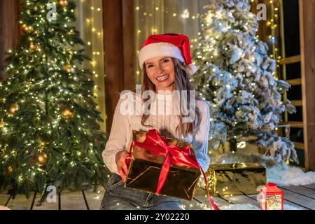 Frau Weihnachtsgeschenk festlich - Eine junge Frau mit Weihnachtsmütze lächelt und öffnet eine Geschenkbox mit rotem Band vor geschmückten Weihnachtsbäumen in A Stockfoto