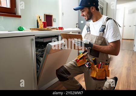 Männlicher Handwerker, der in der Nähe des Geschirrspülers sitzt und auf dem Klemmbrett in der Küche schreibt Stockfoto