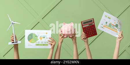 Viele Hände mit Windturbinenmodell, Schweinebank und Diagrammen auf grünem Hintergrund. Konzept der grünen Energie Stockfoto