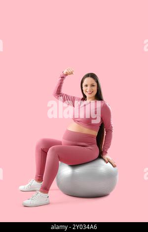 Sportliche schwangere Frau, die Muskeln auf dem Fitball vor rosa Hintergrund zeigt Stockfoto