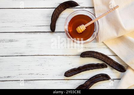 Johannisbrot Honig Konzept, Honig in Glasschüssel mit Johannisbrot Schoten auf weißem Holztisch, gesundes Essen Stockfoto