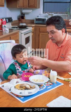 Der spanischer Vater sitzt am Küchentisch und füttert seine 16 Monate alte philippinische Tochter. Modell veröffentlicht MR 1994 ©Bob Daemmrich Stockfoto