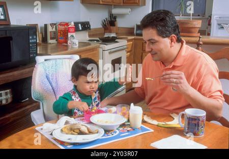 Der spanischer Vater sitzt am Küchentisch und füttert seine 16 Monate alte philippinische Tochter. Modell veröffentlicht MR 1994 ©Bob Daemmrich Stockfoto