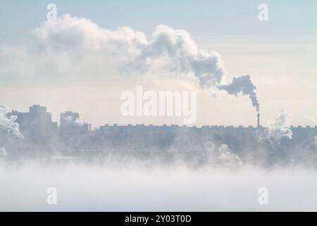 An einem frostigen Wintertag ist die Stadt von Dunst bedeckt. Rauch steigt aus dem Fabrikschornstein auf. Nebel breitet sich im Vordergrund aus. Stockfoto