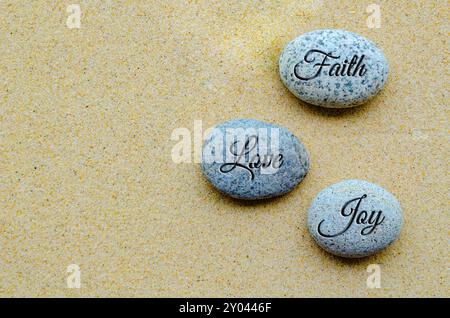 Blick von oben auf den Text von Faith, Love and Joy, der auf Steinen mit Sandhintergrund eingraviert ist. Positives und friedliches Konzept. Stockfoto