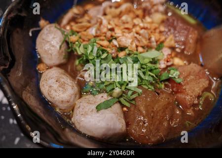 Aus nächster Nähe sehen Sie eine traditionelle thailändische Bootsnudelsuppe, die in einer schwarzen Schüssel serviert wird, mit zarten Schweinefleischbällchen, Schweineschnitzeln und knusprigem gebratenem Knoblauch. Stockfoto