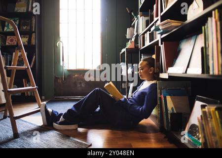 Seitenansicht einer jungen Frau, die Buch liest, während sie sich im Regalregal im Buchladen zurücklehnt Stockfoto