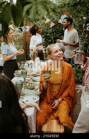 Lächelnde Frau, die ein Glas in der Hand hält und mit einem Freund spricht, während sie am Esstisch sitzt Stockfoto