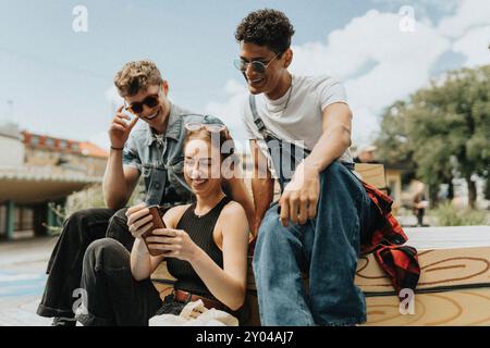 Glückliche junge Frau, die ihr Smartphone mit modischen männlichen Freunden benutzt, während sie auf der Straße sitzt Stockfoto