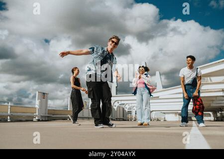 Niedriger Blickwinkel eines glücklichen, modischen jungen Mannes, der mit männlichen und weiblichen Freunden tanzt und auf dem Parkplatz unter Wolken steht Stockfoto