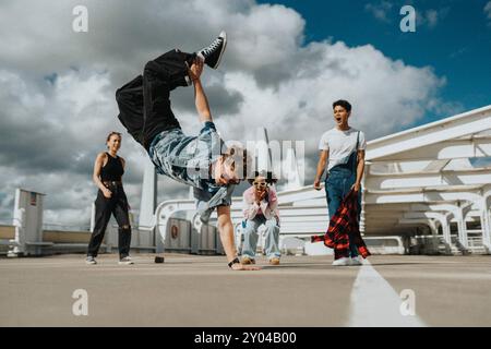 Akrobatischer junger Mann beim Handstand, während männliche und weibliche Freunde auf dem Parkplatz unter bewölktem Himmel jubeln Stockfoto