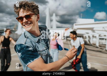 Fröhlicher junger Mann in Sonnenbrille mit tätowiertem Arm, der mit männlichen und weiblichen Freunden auf dem Parkplatz tanzt Stockfoto