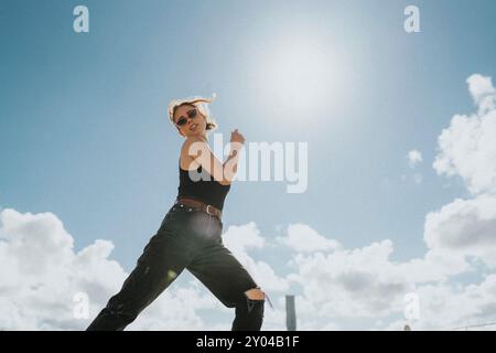 Niedrigwinkelporträt einer jungen Frau in Sonnenbrille tanzend unter bewölktem Himmel an sonnigen Tagen Stockfoto