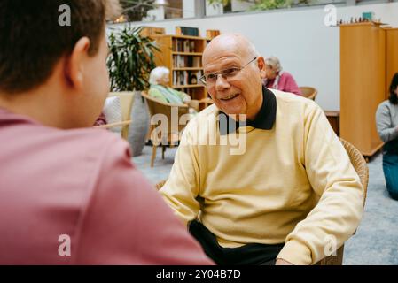 Lächelnder Seniorenmann, der auf Stuhl sitzt und mit männlicher Pflegekraft im Pflegeheim spricht Stockfoto
