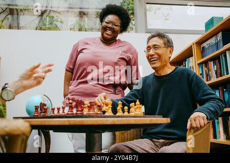 Glückliche Betreuerin, die in der Nähe eines älteren Mannes steht und Schach spielt, während sie im Pflegeheim sitzt Stockfoto