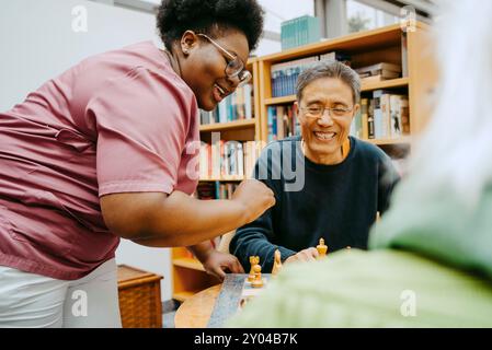 Lächelnde Schwester, die Schach spielt, mit glücklichem Seniorenmann, der im Pflegeheim sitzt Stockfoto
