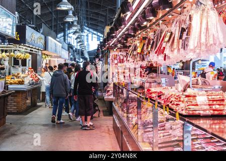 Ein beleuchteter Marktstand bietet eine reiche Auswahl an Schinken- und Fleischprodukten, Kunden im Hintergrund, Mercado de la Boqueria, berühmter Markt auf der Th Stockfoto