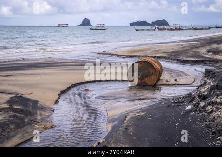 Rostiges Fassöl an einem teilweise schwarzen Strand veranschaulicht die Umweltbelastung durch Ölleckagen. Langschwanzboote im Hintergrund Stockfoto