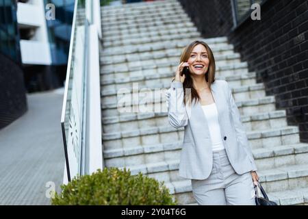 Eine selbstbewusste Frau in einem maßgeschneiderten Anzug schlendert stilvolle Stufen hinunter, lacht über ihr Smartphone und zeigt eine perfekte Mischung aus Professionalität und modernem Li Stockfoto