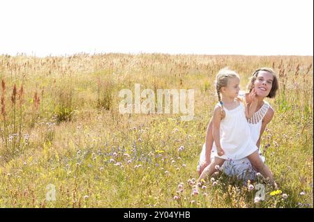 Die schöne junge Mutter und ihre süße kleine Tochter in weißen Sommerkleider sitzen an einem warmen Sommertag auf einer Wiese mit wilden Blumen. Beides Stockfoto