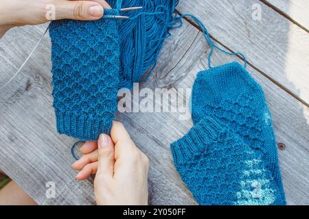 Frau, die blaue Strickarbeit auf grauem hölzernem Hintergrund hält. Strickmädchen im Garten. Blaues Stricken in Mädchenhänden auf Holztisch. Stockfoto