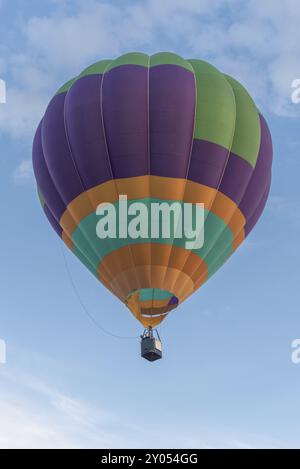 Bunt gefärbter Heißluftballon, der an einem klaren Tag in den Himmel fliegt, von mehreren Personen gesteuert. Die Hintergrundlandschaft zeigt einen hellblauen Himmel und einige s Stockfoto